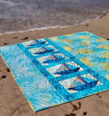 Top US quilting blog & shop, Kate Colleran Designs, shares about a sailboat quilt pattern Sail Away and a cute batik crib quilt! Image is a of a blue and green sailboat quilt on the sand at the beach