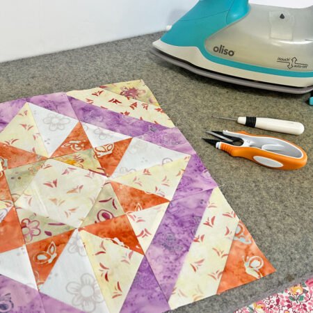 Top US quilting blog and shop, Kate Colleran Designs, reveals her February Remix Quilt Block! Image is of a star quilt block in green, yellow, orange and purple with an iron and snips. 