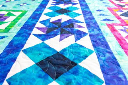 Top US quilting blog and shop, Kate Colleran Designs, shares about her new block of the month quilt using Aura fabrics from QT! Image is a closeup of quilt blocks in mottled blue and purple fabrics.