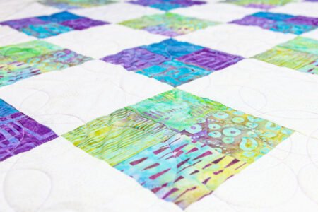 Top US quilting blog and shop, Kate Colleran Designs, shares about a batik line Fairy Floss and her quilt pattern Four Square. Image is a closeup of a quilt using squares of blue, purple and green batiks.