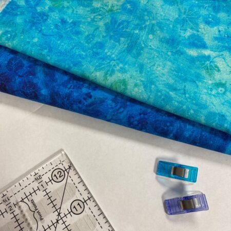 Top US quilting blog and shop, Kate Colleran Designs, shares a free quilt block pattern and shares about an upcoming QAL! Image is of 2 blue and 1 white fabric along with binding clips and part of a ruler.