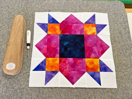 Top US quilting blog and shop, Kate Colleran Designs, shares tips in the Allure BOM Block 7 post. Image is a quilt block in bright purple, magenta pink, orange and white on a grey wall pressing mat with a brown wooden clapper and white seam ripper next to the block.