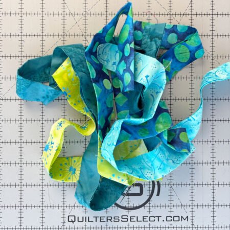 Top US quilting blog and shop, Kate Colleran Designs, shares about the Bias Tape Challenge and her project that she created. Image is of blue and green batik strips in a pile on a cutting mat.