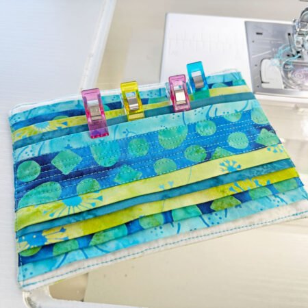 Top US quilting blog and shop, Kate Colleran Designs, shares about the Bias Tape Challenge and her project that she created. Image is a closeup of blue and green batik strips sewing together with binding clips attached to the folds.