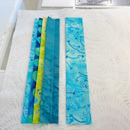 Top US quilting blog and shop, Kate Colleran Designs, shares about the Bias Tape Challenge and her project that she created. Image is a closeup of blue and green batik strips being sewn to a white back.