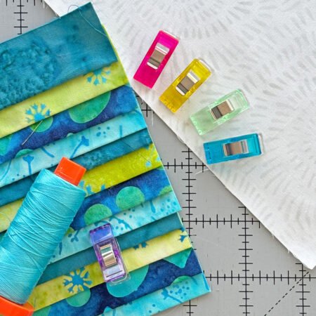 Top US quilting blog and shop, Kate Colleran Designs, shares about the Bias Tape Challenge and her project that she created. Image is a closeup of blue and green batik strips, blue thread spool and colorful binding clips.