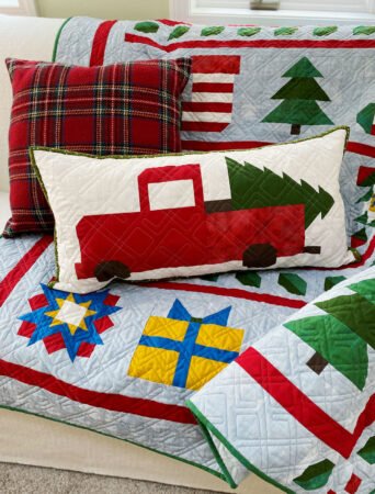 Top US quilting blog and shop, Kate Colleran Designs, shares about the Jingle Bell Pines Block of the Month quilt Block Party. Image is a holiday quilt with a medium blue background and pieced holiday blocks in red, green, white and gold draped on a couch with a holiday truck block pillow.