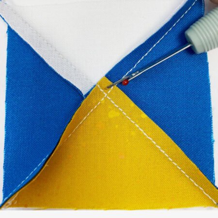 Top US quilting blog and shop, Kate Colleran Designs, shares about the Jingle Bell Pines Block Party Block 7 Rose Star. Image is the back of a quarter square triangle unit in blue, white and gold with a seam ripper to show which thread to clip.
