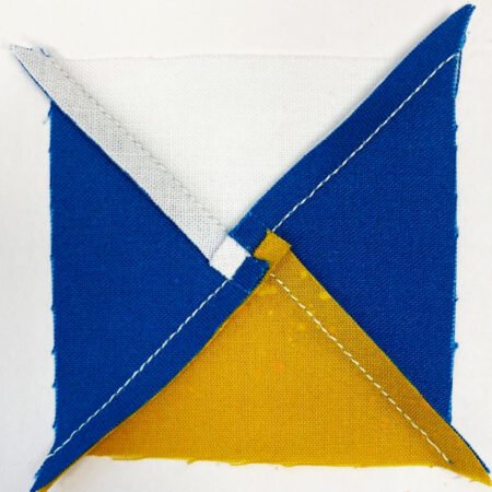 Top US quilting blog and shop, Kate Colleran Designs, shares about the Jingle Bell Pines Block Party Block 7 Rose Star. Image is the back of a quarter square triangle unit in blue, white and gold with swirled seams.