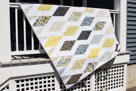 Top US quilting blog and shop, Kate Colleran Designs, shares about her quilt Gemstone and the quilt remake in Blissful Blooms. Image is a bold geometric quilt in black, yellow, grey and white draped on a porch railing.