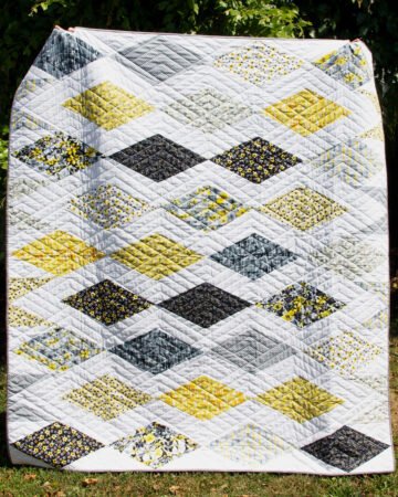 Top US quilting blog and shop, Kate Colleran Designs, shares about her modern FQ friendly quilt Gemstone made in QT Fabrics. Image is a modern diamond quilt in yellow, grey, black and white modern fabrics with florals, leaves, mini prints and graphic brushes of color .