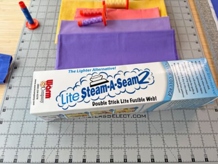 a box of Lite Steam a Seam 2 with the stack of fabrics and threads behind it in purples, coral and yellow