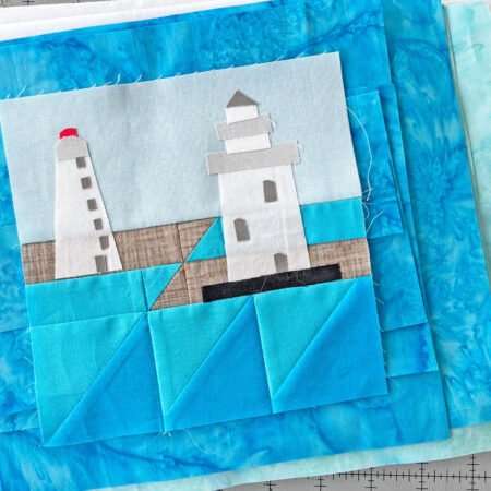 stack of aqua and blue batik fabrics with appliqués lighthouse quilt block on top of the pile 