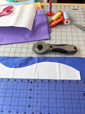  royal blue fabric with a wavy shape cut out from white fusible with sewing supplies surrounding the fabric.
