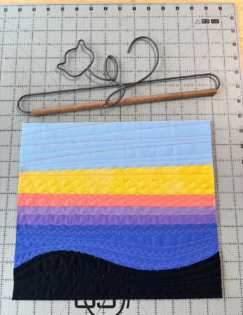  a quilt top quilted and on a cutting table with a decorative wire and wood hanger above it. Quilt is lines of colors- light blue, yellow, coal, lavender, purple, royal blue and black. 