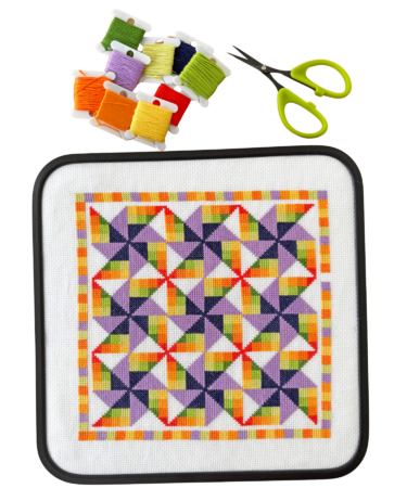 cross stitch project showing quilt blocks in purple, dark blue, greens, yellow, orange and red with bobbins of thread and scissors